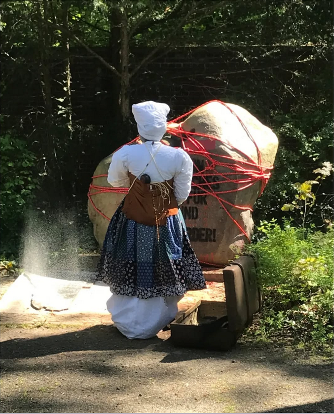 Frau in traditionellem namibischem Gewand mit weißem Turban steht mit dem Rücken zum Stein mit der Inschrift. Der Stein ist mit Schnur umwickelt.
