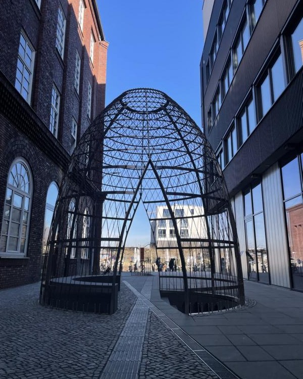 Gitterartiges, eiförmiges Gebilde aus Metall zwischen zwei Häusern. Es hat zwei offene Bereiche, durch die man hindurchgehen kann.