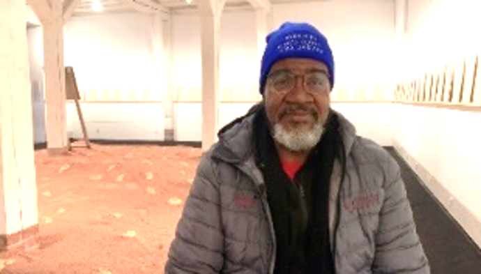 Ein Mann sitzt frontal zur Kamera. Im Hintergrund sieht man eine Installation mit rotem Sand. An den Wänden befinden sich Texttafeln.