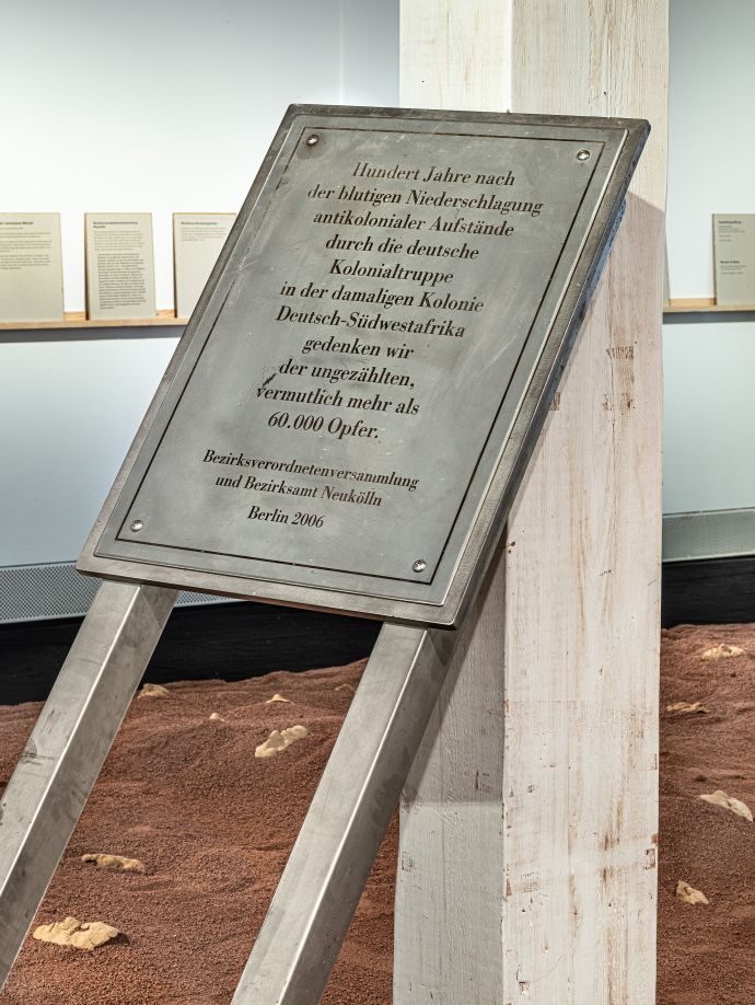 Metallplatte mit Inschrift ist gegen eine weiße Säule gelehnt. Im Hintergrund ist roter Sand mit zum Teil vergrabenen Tonmasken menschlicher Gesichter zu sehen.