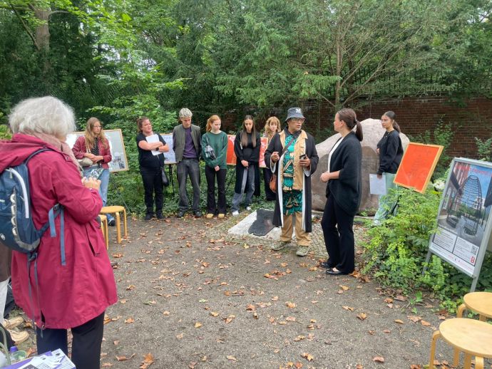 Micohael Küppers-Adebisi und Bezirksstadträtin Janine Wolter im Gespräch (Foto: FB MSE 2025) Eine Gruppe von Personen unterschiedlichen Alters und Geschlechts steht im Halbkreis um den sog. Hererostein. In der Mitte stehen ein Mann und eine Frau und unterhalten sich.