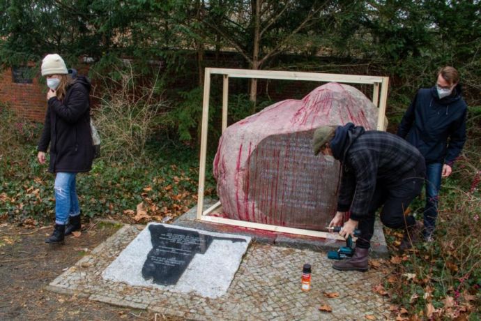 Ein großer Stein mit Inschrift wird durch drei Personen mit FFP2-Masken mit einem Holzrahmen versehen. 