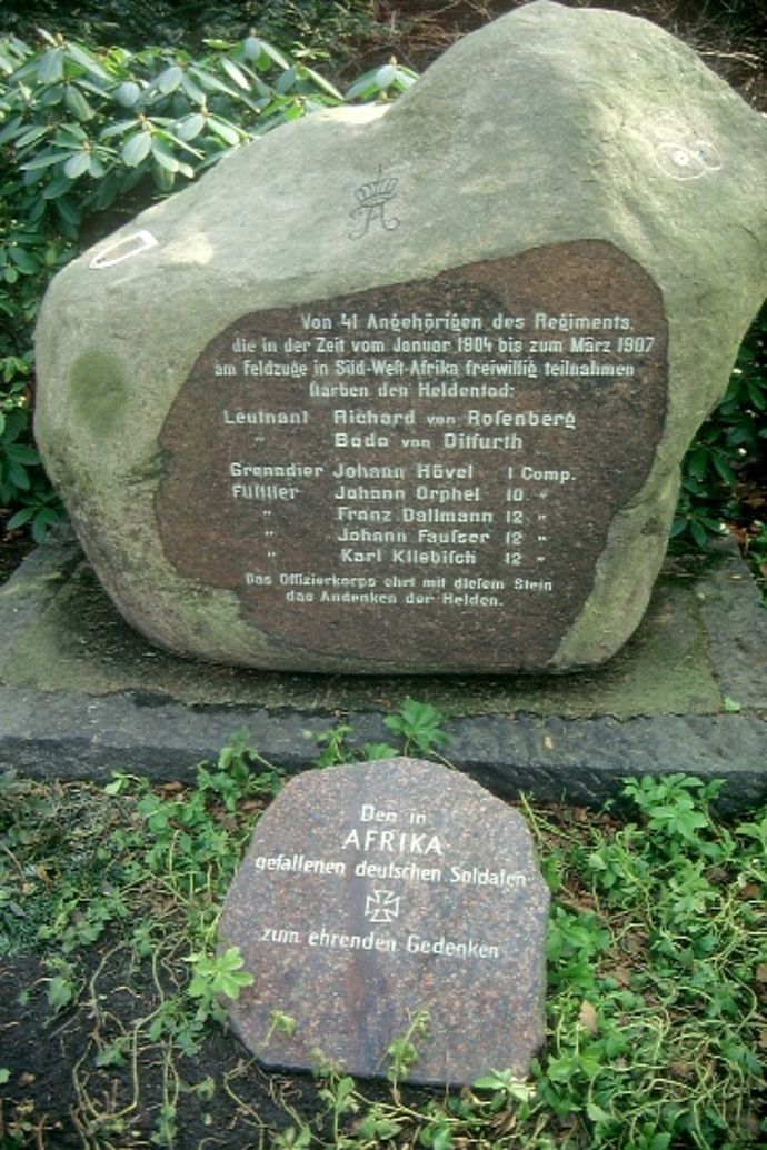 vor einem großen Stein mit Inschrift liegt ein kleinerer Stein mit Inschrift. Drum herum sind grüne Pflanzen zu sehen.