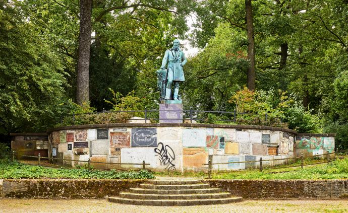 Statue eines Mannes auf einem Sockel aus verschiedenen Steinen. Vorne führt eine Treppe hoch. Jahn Denkmal in der Hasenheide, Link zur Ausstellung