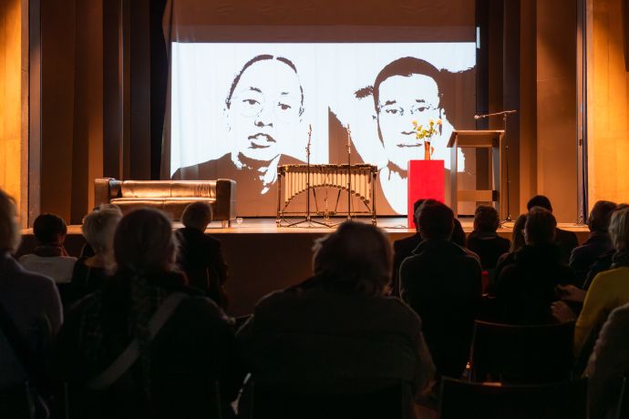 Eine Bühne mit einem Musikinstrument, einem Sofa und einem Rednerpult. Im Hintergrund ein Bild in Schwarz-Weiß, dass 2 Menschen zeigt, die direkt in die Kamera schauen.