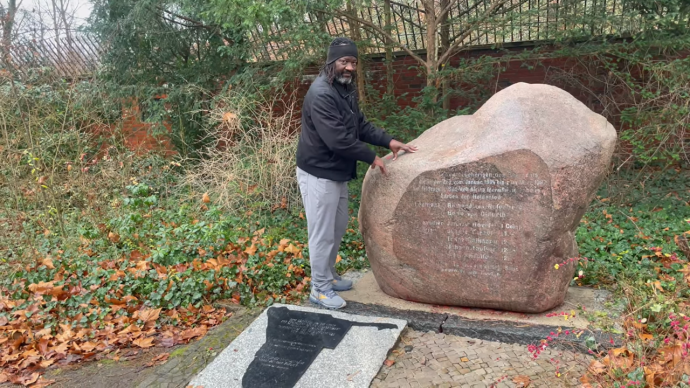 Ein Mann steht an dem Stein mit der Inschrift. Er hat beide Hände auf den Stein gestützt.