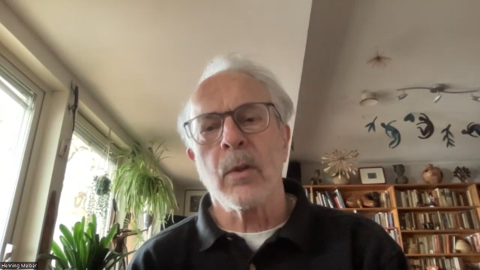 Ein Mann sitzt frontal zur Kamera, im Hintergrund ist ein Arbeitszimmer mit Bücherregal und Pflanzen zu sehen.