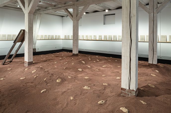 Ausstellungsraum. In der Mitte ist eine Landschaft aus rotem Sand, in die Tonmasken menschlicher Gesichter eingegraben ist. Links steht ein metallenes Schild an eine Säule gelehnt, die Schrift zeigt in eine andere Richtung. An den Wänden läuft ein schmales Holzregal, auf dem Texttafeln in DinA4 Format stehen,.