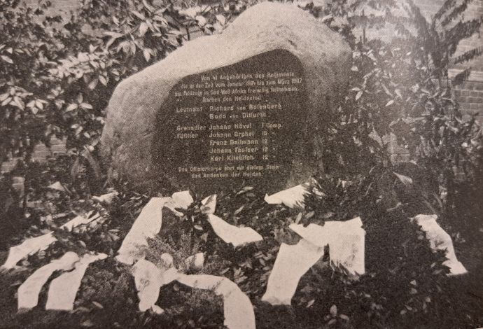 schwarz weiß Fotografie eines großen Steins mit Inschrift. Um den Stein herum sind Pflanzen. Vor dem Stein liegen Blumenkränze.