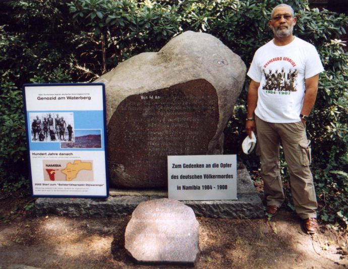 neben dem Stein mit Inschrift stehen zwei Plakate. Rechts vom Stein steht ein Mann mit einem T-Shirt, auf dem Ovaherero Genocide steht.