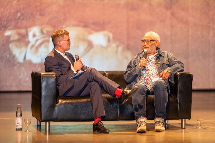 Zwei Personen sitzen auf einem Sofa und unterhalten sich. Sie halten Mikrofone. Im Hintergrund ist ein Bild einer menschlichen Tonmaske auf einer Leinwand.
