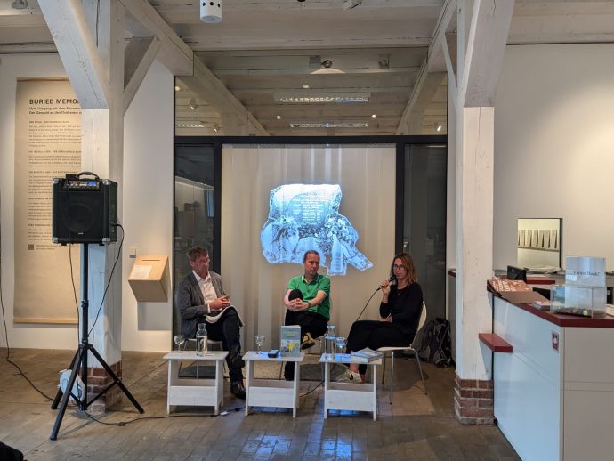 zwei Männer und eine Frau sitzen gemeinsam vor einem Plastikvorhang, auf den ein Bild projeziert wird. Sie diskutieren miteinander.