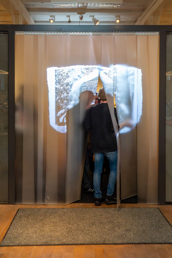 Eien Person geht durch einen Vorhang, der aus breiten Plastiklamellen besteht. Auf den Vorhang wird ein bild projeziert.