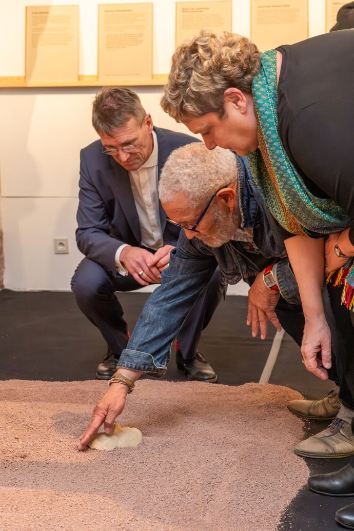 Zwei Männer und eine Frau beugen sich zu einem Sandhaufen. Der Mann in der Mitte vergräbt gerade eine Tonmaske eines menschlichen Gesichts.