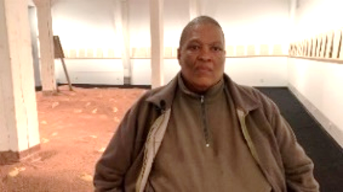 Ein Mann sitzt frontal zur Kamera. Im Hintergrund sieht man eine Installation mit rotem Sand. An den Wänden befinden sich Texttafeln.