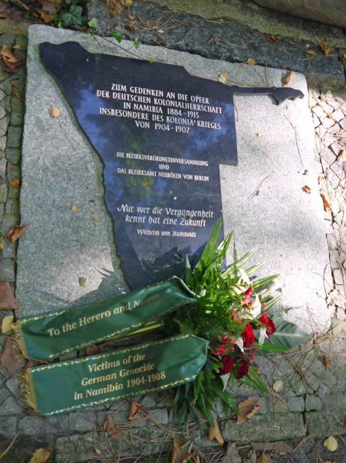schwarze Gedenkplatte in Form des Landes Namibia mit Blumen daneben.