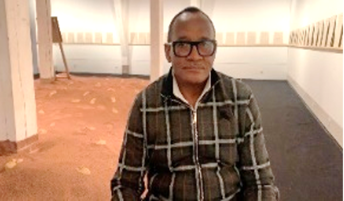 Ein Mann sitzt frontal zur Kamera. Im Hintergrund sieht man eine Installation mit rotem Sand. An den Wänden befinden sich Texttafeln.