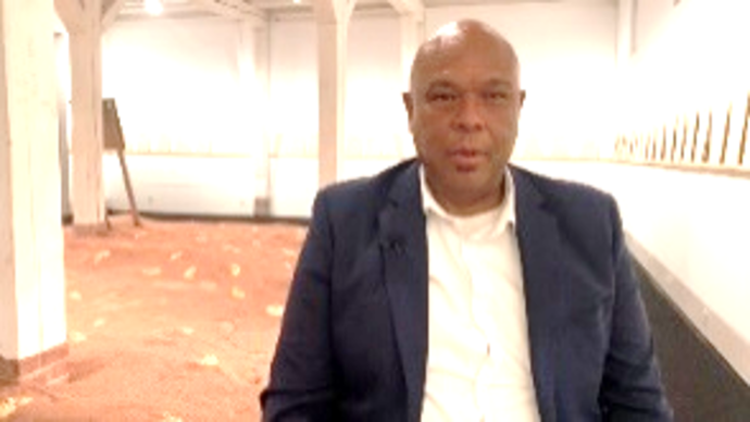 Ein Mann in anzug sitzt frontal vor der Kamera. Im Hintergrund zu sehen ist ein weißer Raum mit einer Installation aus rotem Sand im Hintergrund. An den Wänden sind Textafeln in DinA4 Format.