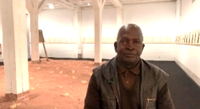 Ein Mann sitzt frontal zur Kamera. Im Hintergrund sieht man eine Installation mit rotem Sand. An den Wänden befinden sich Texttafeln.