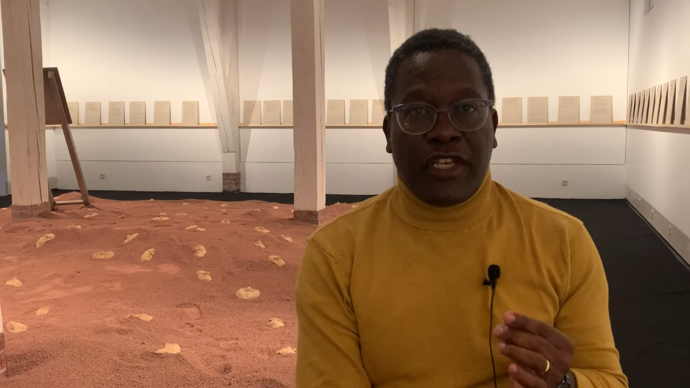 Ein Mann sitzt frontal zur Kamera. Im Hintergrund sieht man eine Installation mit rotem Sand. An den Wänden befinden sich Texttafeln.